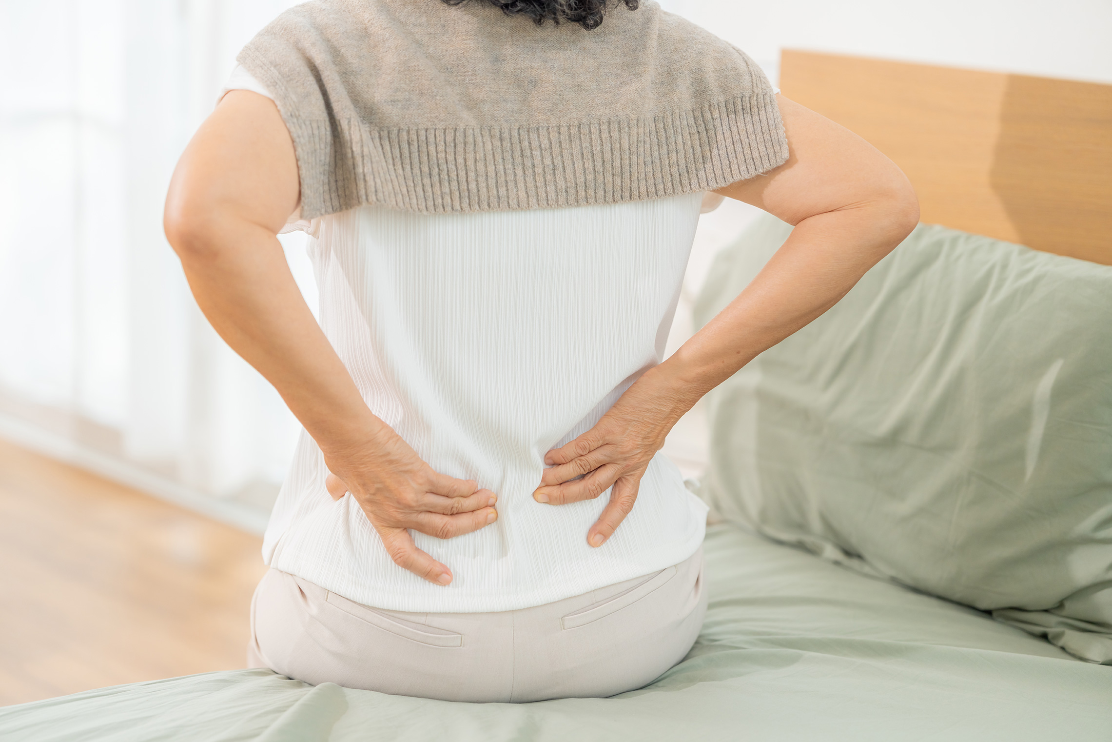 Woman sitting on the bed experiencing back stiffness and discomfort after waking.